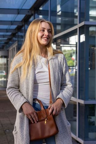 Smiling woman standing outdoors holding brown leather crossbody handbag under glass canopy