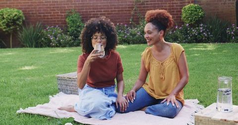 Two women enjoying picnic on green lawn sharing refreshing water and laughing together