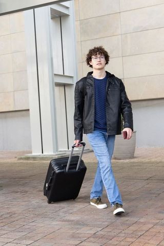 Young man walking with rolling suitcase and checking smartphone outside beige building