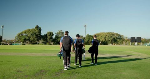 Cricket teammates walking to pitch carrying bats helmets pads kit bags on sunny outfield