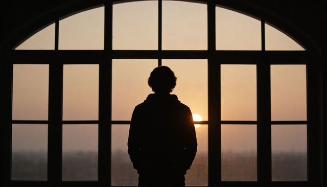 Solitary silhouette watching sunset through arched window at dusk