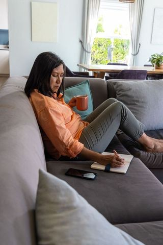 Mature woman writing journal while sipping coffee on gray sofa with smartphone nearby