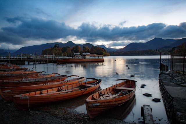 Empty wooden boats anchored on a calm lake against a backdrop of mountains and a cloudy sunset sky. Ideal for use in travel brochures, nature magazines, and websites promoting relaxation and outdoor adventures. Highlights peaceful moments in nature and evokes feelings of tranquility and escape.