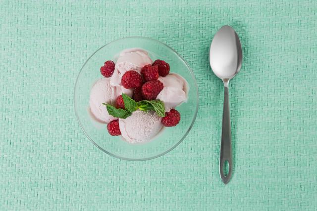 Raspberry ice cream served in a glass bowl, topped with fresh raspberries and mint leaves, placed on a textured green background. A spoon is placed beside the bowl. Ideal for use in food blogs, dessert menus, summer-themed promotions, and social media posts highlighting refreshing treats.