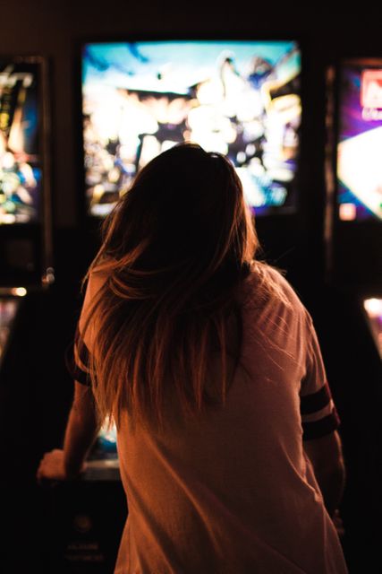 A woman with long hair in a casual shirt enjoying a game of pinball in a dimly lit arcade. The lighting focuses on the pinball machine, creating an engaging and immersive gaming atmosphere. Ideal for use in articles about gaming culture, recreational activities, or retro entertainment. This could also be useful in promotional materials for arcade venues or articles highlighting nostalgic experiences.