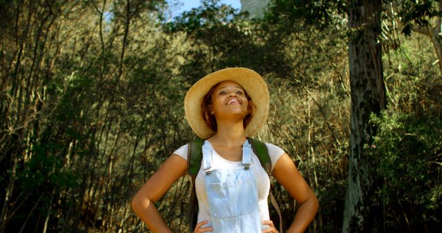 Young woman enjoying a hike in the forest, wearing a straw hat and a backpack. Ideal for use in content about outdoor activities, nature explorations, adventure, and travel. Suitable for promoting hiking gear, travel destinations, and wellness blogs.