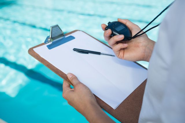 Swim coach holding a stopwatch and clipboard at poolside, focusing on timing and training. Ideal for use in articles about swimming training, coaching techniques, sports timing, and aquatic sports preparation.