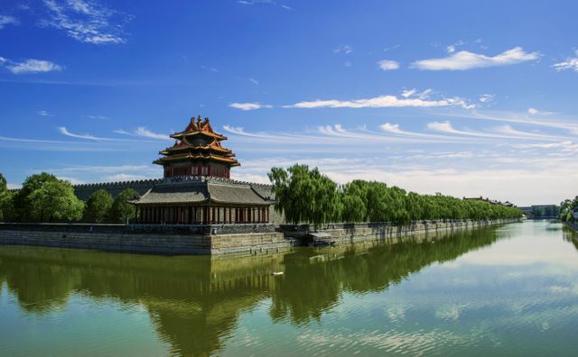 Elegant image showcasing traditional Chinese pavilion situated beside a tranquil river with lush green trees lining the water and clear blue skies above. Ideal for travel brochures, cultural studies, history features, and nature presentations.