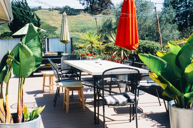 Sunny outdoor patio featuring a table with chairs arranged around it, surrounded by lush plants and a vibrant red umbrella. Ideal for backyard gatherings, barbecue parties, outdoor dining, or relaxation in a garden setting.