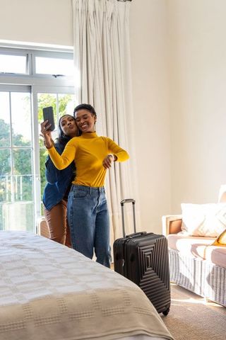 Diverse friends taking selfie in sunlit hotel room with suitcase and balcony view
