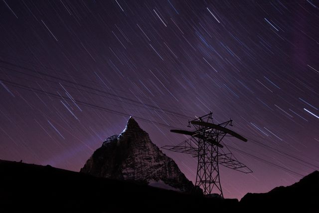 Capturing star trails above mountain peak creates a mesmerizing night sky scene. The silhouette of the mountain and an electric tower contrasts against the purple hues of the night, with power lines adding an element of human connection. Perfect for use in nature, travel, and adventure blogs, educational material on astrophotography, and visually stunning backgrounds.