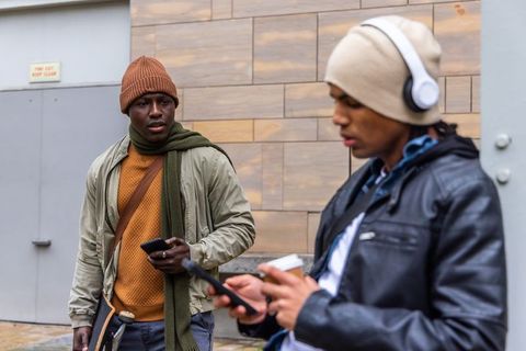 Diverse young men standing on city sidewalk wearing beanies holding phones and skateboard