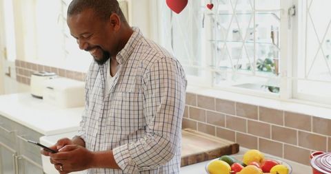 Happy Man in Kitchen Texting on Smartphone with Fresh Fruit