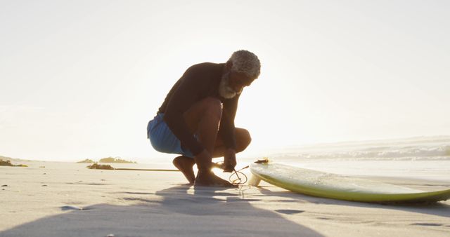 Elderly man adjusting his surfboard leash on the beach during sunset, showcasing active lifestyle and love for surfing. Ideal for use in articles on senior fitness, outdoor activities, surfing culture, beach vacations, and healthy aging.