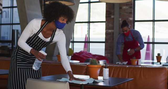 Restaurant staff are thoroughly cleaning tables while wearing masks, emphasizing hygiene and safety during the pandemic. This visual is suitable for materials related to COVID-19 safety protocols, workplace hygiene, and the food service industry's response to health regulations.