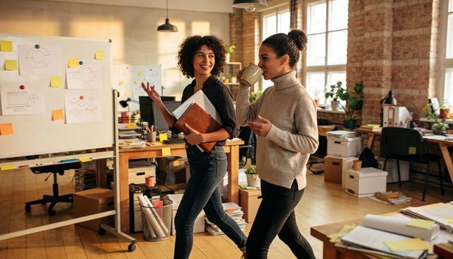 Women walking in open-plan studio discussing ideas, sipping coffee, carrying notebooks