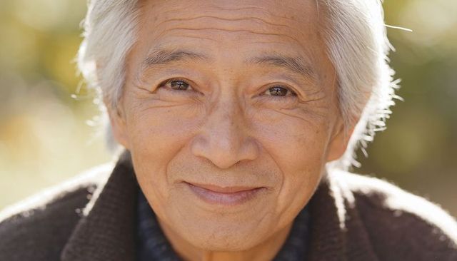 Sunlit senior asian man smiling at camera in park during golden hour — gentle portrait