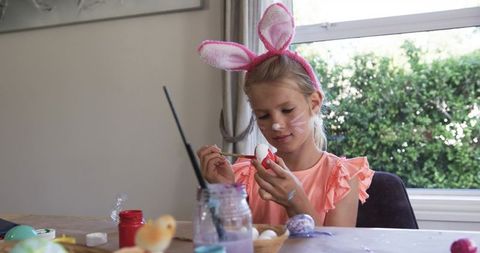 Girl wearing bunny ears painting easter eggs at table, spring craft and family fun