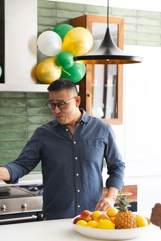 Senior asian man standing in kitchen wearing blue shirt and glasses arranging fruit bowl