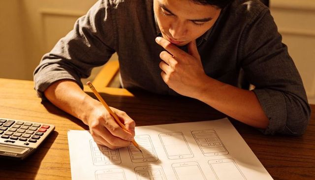 Leaning designer sketching mobile app wireframes on paper with pencil and calculator