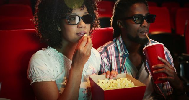 Couple sitting in theatre, wearing 3D glasses, enjoying movie with popcorn and soda. Perfect for concepts related to entertainment, friendship, date night, leisure activities, cinema experiences, youthful fun, or movie-related promotions.