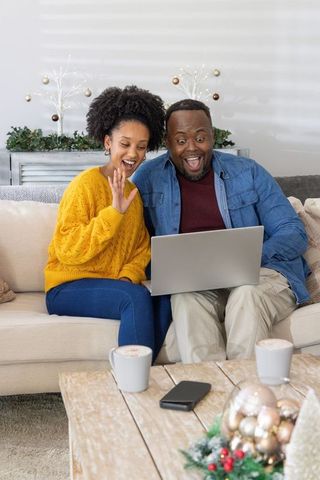 African american couple waving during joyful holiday video call on laptop in cozy living room
