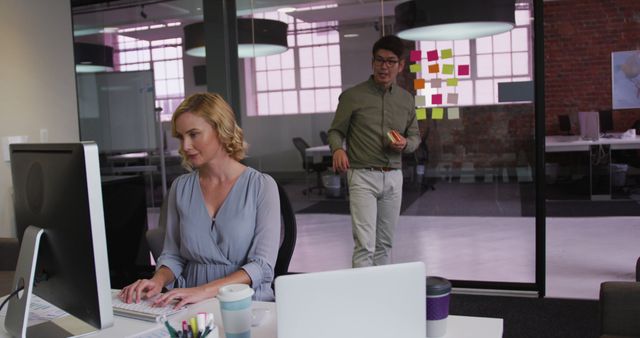 Woman Working at Desk in Modern Office While Colleague Stands in Background with Snack - Download Free Stock Images Pikwizard.com