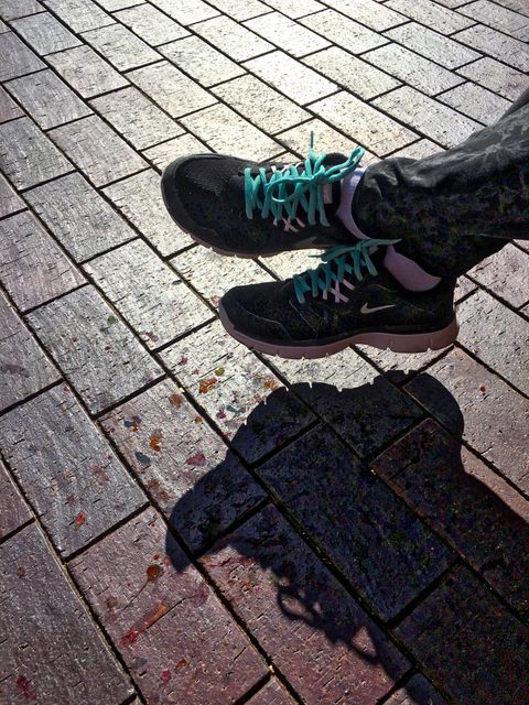 This image shows a pair of sport shoes with teal laces walking on a brick pavement in an urban outdoor setting. Shadows add dimension and highlight the street textures. Ideal for promoting streetwear fashion, outdoor activities, urban lifestyles, or footwear brands.
