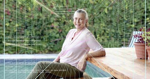 Smiling senior woman relaxing poolside on patio leaning on wooden table with plants