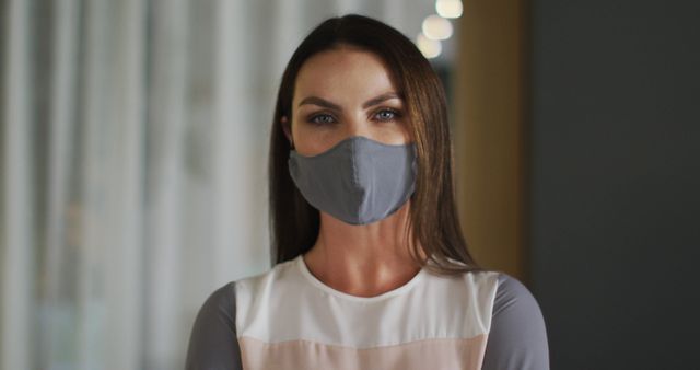 Woman with long dark hair wearing gray protective face mask standing indoors in professional environment. Could be used for articles or advertisements related to health, pandemic safety, and protective measures during COVID-19.