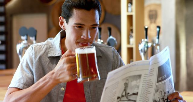 A young Asian man enjoys a pint of beer while reading a newspaper at a bar, with copy space. His relaxed posture and the casual setting suggest a leisurely break in his day.