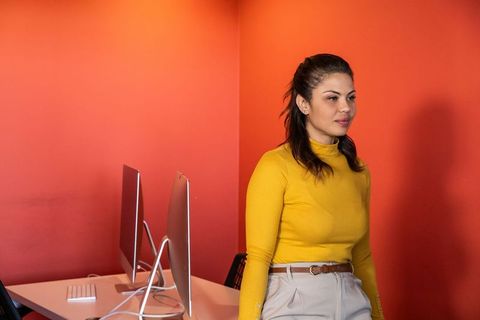 Young woman standing in modern office with dual monitors, wearing mustard sweater and beige trousers