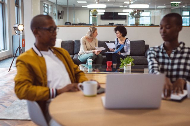 Diverse group of creative professionals collaborating in a modern office lounge area. Two women are engaged in a discussion on a sofa, while two men are working on laptops at a nearby table. Ideal for depicting teamwork, modern work environments, and casual business settings.