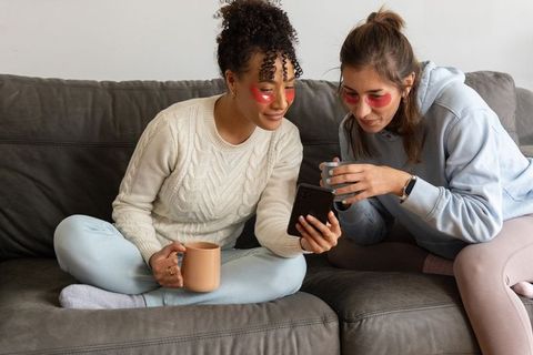 Diverse female friends relaxing on sofa sharing smartphone and under-eye gel patches