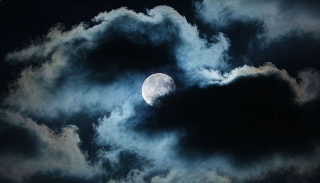 Nearly-full moon peeking through dramatic cloud halo casting cool blue nocturnal glow