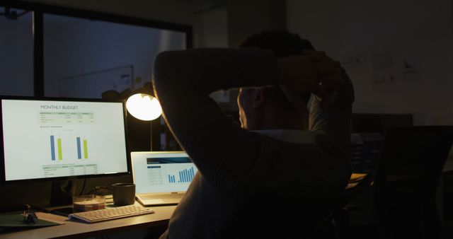 Person in a dark office working late with budget analysis shown on computer screens performing financial tasks, diligence. Ideal for illustrating workplace stress, overworking scenarios, financial industry, or articles related to workaholism and productivity.