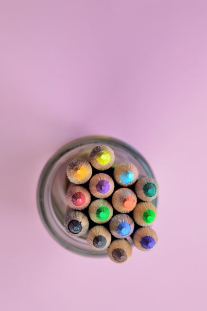 A collection of sharpened color pencils neatly arranged in a glass jar viewed from the top against a pink background. Vibrant and ready for drawing or coloring activities, perfect for advertising educational tools, creative workshops, or art supply products.