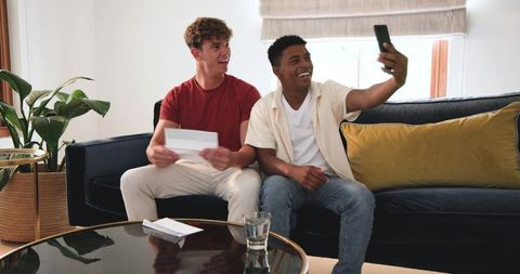 Diverse friends taking selfie on modern navy sofa while opening greeting card and laughing