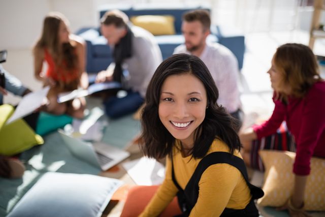 Smiling businesswoman sitting in a modern office with colleagues in the background. Ideal for depicting teamwork, collaboration, and a positive work environment. Suitable for corporate websites, business presentations, and promotional materials highlighting professional success and diversity.