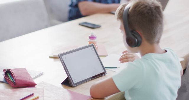 Young boy engaged in online learning at home, using a tablet and headphones. Ideal for illustrating home study environments, modern education, digital learning tools, and homeschooling setups.