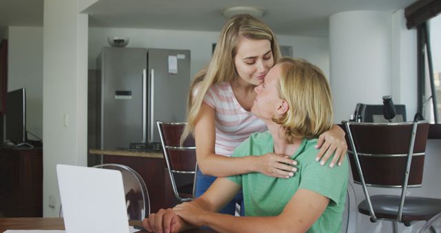This image shows a young couple sharing a tender moment in a modern kitchen. The scene includes a woman embracing and kissing a man who is seated in front of a laptop. This image can be used for themes related to romance, daily life, home, and modern technology.
