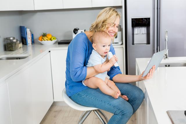 Mother holding baby boy while using digital tablet in a modern kitchen. Ideal for content related to parenting, family life, technology use in daily routines, and modern home settings. Perfect for blogs, articles, and advertisements focusing on family bonding, tech-savvy parenting, and home lifestyle.