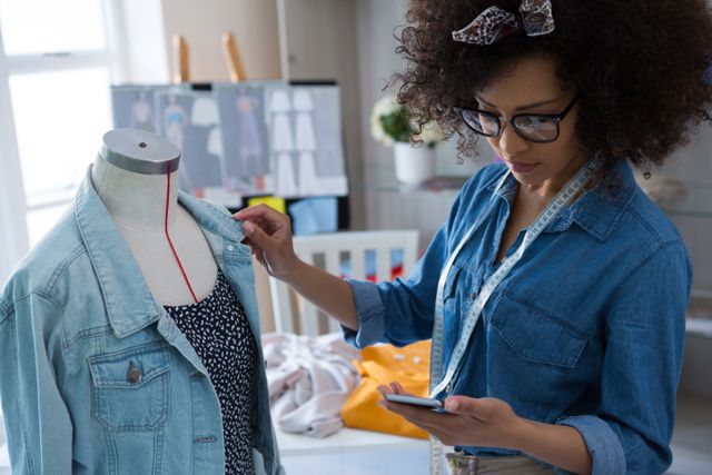 Female fashion designer using mobile phone while designing a dress at home. She is adjusting a denim jacket on a mannequin and has a measuring tape around her neck. Ideal for content related to fashion design, home-based businesses, creativity, and the fashion industry.