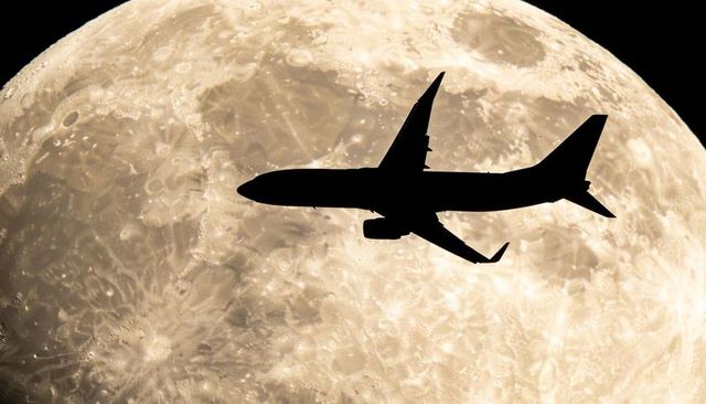 Jet airliner silhouette flying across full moon with detailed lunar surface