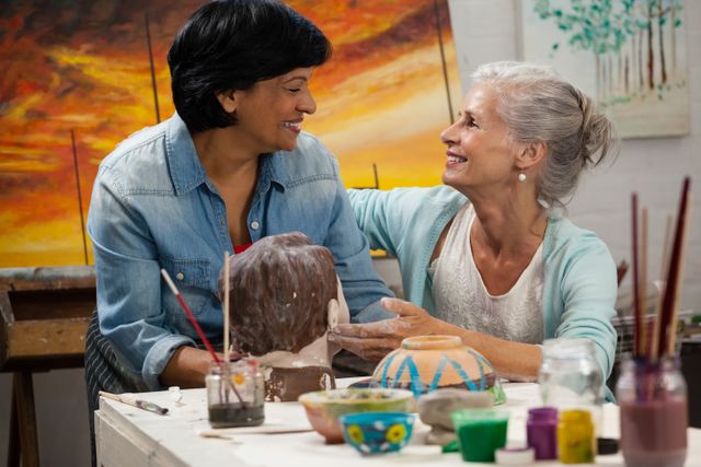 Two women, one senior and one adult, are enjoying a pottery class together. They are painting a sculpture and smiling at each other, showcasing a moment of bonding and creativity. This image can be used for promoting art classes, workshops, senior activities, or community events focused on creativity and friendship.