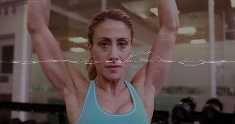 Focused mid-adult woman lifting arms in gym with waveform overlay for fitness tracking