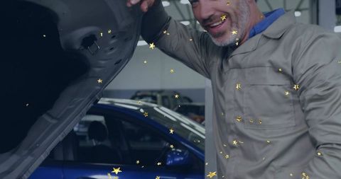 Smiling mechanic leaning under hood wearing coveralls beside blue sedan with gold stars