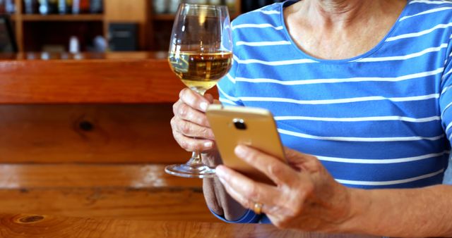 Senior woman holding wine glass and using smartphone in rustic cafe. Perfect for illustrating themes like leisure activities, technology usage among seniors, smartphone communication, and relaxation. Ideal for blogs, articles on modern senior lifestyles, and advertisements featuring older adults.