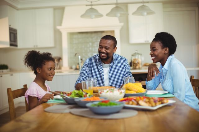 A joyful African American family is enjoying a meal together at home. The family members are sitting at a dining table filled with various dishes, including vegetables, rice, and corn on the cob. This image can be used for themes related to family bonding, home life, happiness, and healthy eating. It is perfect for advertisements, blogs, or articles focused on family values, parenting tips, or kitchen and home decor.