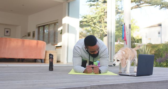 African american man exercising using laptop on sunny terrace with dog. Lifestyle, exercise, fitness, healthy lifestyle, nature, communication, animal and domestic life, unaltered.